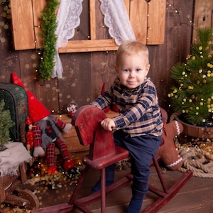 Könnte beinhalten: Ein junges Kind sitzt auf einem roten Schaukelpferd aus Holz. Das Kind trägt ein kariertes Hemd und eine marineblaue Hose. Der Hintergrund zeigt eine rustikale Holzwand und Weihnachtsdekorationen.