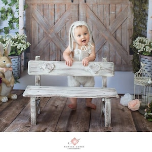 Peut inclure: Un bébé portant un costume de lapin blanc est assis sur un banc en bois blanc avec des détails en forme de cœur. Le banc est devant une porte en bois rustique avec un fond blanc.