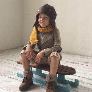 Può includere: Un bambino con un cappello da aviatore marrone, una sciarpa giallo senape e un cappotto beige è seduto su una slitta di legno con pattini blu. Il bambino indossa anche stivali marroni e scaldamuscoli a maglia. La slitta è su un pavimento di legno bianco.