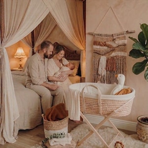 May include: A cozy nursery scene with a woven bassinet on a wooden stand, a plush rug, and a couple holding a baby. A woven wall hanging and a lamp add to the warm, neutral tones of the room. A stuffed lion and a goose are in the scene.