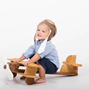 Può includere: Un bambino piccolo con un cappello da aviatore marrone con paraorecchie bianche è seduto su un aereo di legno giocattolo. L'aereo ha un'elica bianca ed è dipinto di marrone.