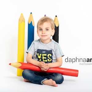 Può includere: Un bambino piccolo è seduto a gambe incrociate, tenendo un grande matita rossa. Indossa una maglietta grigia con le parole "Say Yes" stampate su di essa. Dietro di lui ci sono quattro grandi matite gialle, blu, viola e nere.