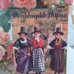 Puede incluir: Una postal vintage con tres mujeres vestidas con trajes tradicionales galeses. Las mujeres están de pie en un campo con un muro de piedra en el fondo. La postal tiene el texto "Penblwydd Hapus" escrito en ella, que en galés significa "Feliz cumpleaños".