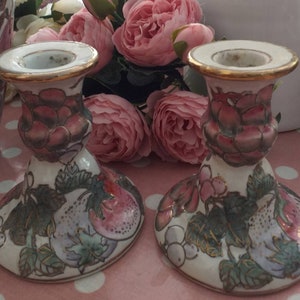 May include: A pair of white ceramic candle holders with a gold rim. The holders are decorated with a floral and fruit design in shades of pink, green, and purple.