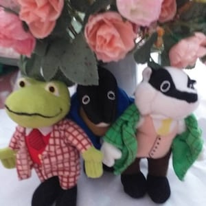 May include: Three plush toys: a green frog wearing a red and white checkered jacket, a black and white badger wearing a green jacket, and a black and white otter wearing a blue jacket.
