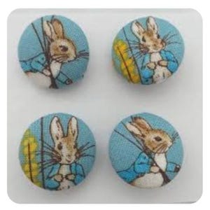Puede incluir: Cuatro botones de tela azul con un diseño de Peter Rabbit. Cada botón presenta un conejo blanco con una chaqueta azul y sosteniendo una zanahoria. El fondo es de color azul claro con acentos amarillos.