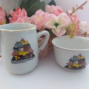 Puede incluir: Un juego de jarra de crema y azucarero de cerámica blanca con ribete dorado. El juego presenta una imagen de dibujos animados de Tom y Jerry con el texto "Tom & Jerry".