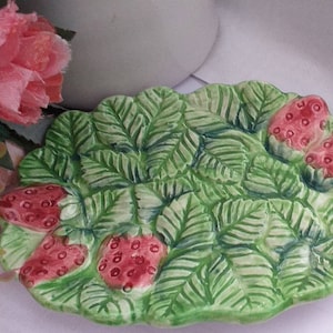 May include: A ceramic dish shaped like a strawberry leaf with four red strawberries. The dish is green with a light green and white pattern.
