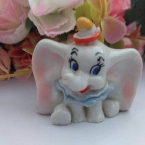 Puede incluir: Una pequeña figurita de cerámica de Dumbo el elefante de la película animada de Disney Dumbo. La figurita está pintada de blanco con orejas rosas, detalles azules y rojos y ojos negros.