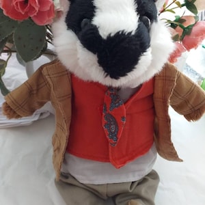May include: A plush badger wearing a brown plaid jacket, a red waistcoat with a paisley tie, a white shirt, and beige trousers. The badger is sitting on a white surface.