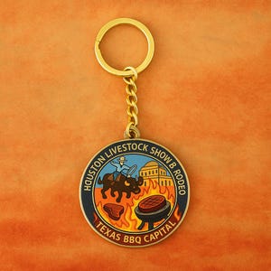 K&ouml;nnte beinhalten: Ein runder Schl&uuml;sselanh&auml;nger aus goldfarbenem Metall mit einem farbigen Emaille-Design. Die Mitte zeigt einen Cowboy auf einem Stier, ein Geb&auml;ude und einen Grill mit einem Steak &uuml;ber Flammen. Der Text lautet HOUSTON LIVESTOCK SHOW & RODEO und TEXAS BBQ CAPITAL.