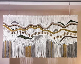 May include: A white macrame wall hanging with brown, gray, and yellow accents. The hanging features a wavy pattern with fringe at the bottom.