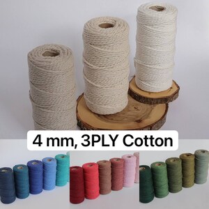 Peut inclure: Plusieurs bobines de corde en coton 4 mm, 3 fils, de différentes couleurs. L'image présente de grandes bobines de corde naturelle et blanc cassé, ainsi que de plus petites bobines bleues, rouges et vertes. Le texte "4 mm, 3PLY Cotton" est visible.