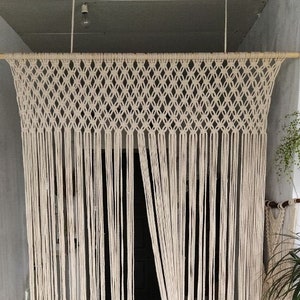 May include: A child stands behind a macrame curtain made of natural cotton rope. The curtain is hung from the ceiling and has a woven pattern. The child is wearing a blue jumpsuit and is looking up.