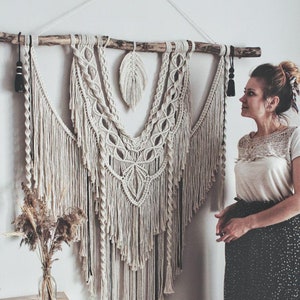 Könnte beinhalten: Ein großer Makramee-Wandbehang mit einem Holzstab oben. Der Makramee ist aus naturfarbenem Garn gefertigt und hat ein Federdesign in der Mitte. Der Wandbehang hängt an einer weißen Wand und eine Person steht rechts davon.