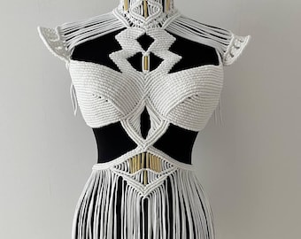 May include: A white macrame dress displayed on a black mannequin. The dress features a high collar, shoulder accents, and a fringed skirt. Gold-colored accents are incorporated into the design, adding a touch of elegance to the handmade garment.