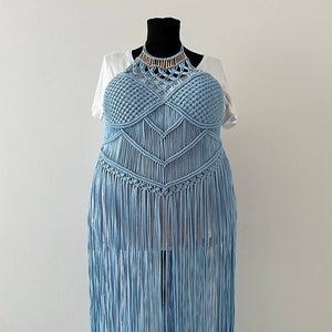 May include: A light blue crochet top with a halter neckline and fringe detailing. The top is made with a macrame technique and features a gold beaded necklace.