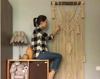 May include: A woman is hanging a large, beige macrame wall hanging on a wall. The wall hanging is made of cotton cord and has a geometric design. The woman is wearing a plaid shirt and jeans. The wall hanging is hanging on a wooden dowel rod.