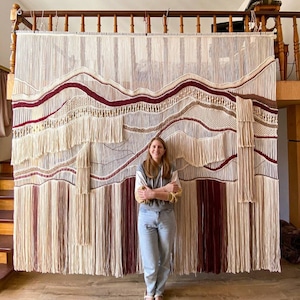May include: A large wall hanging made of woven yarn with a variety of textures and colors, including white, beige, and burgundy. The hanging is made up of several layers of yarn, creating a three-dimensional effect. A person is standing in front of the hanging, providing a sense of scale.