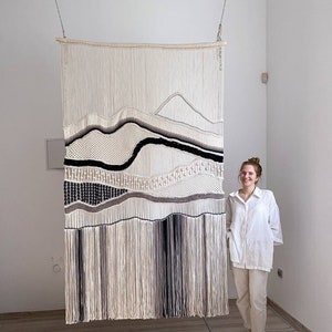 May include: A large, white macrame wall hanging with black and brown accents. The hanging features a geometric design with a wavy pattern. The hanging is suspended from the ceiling by two chains.