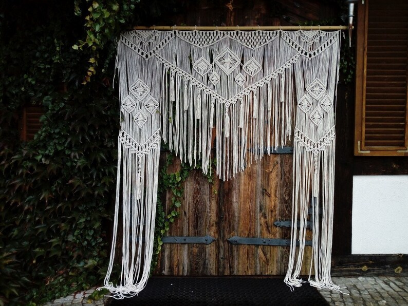 May include: A white macrame wall hanging featuring geometric diamond designs and long fringe. The handmade textile is suspended from a wooden rod, creating a bohemian decorative element. The macrame hangs in front of a rustic wooden door.