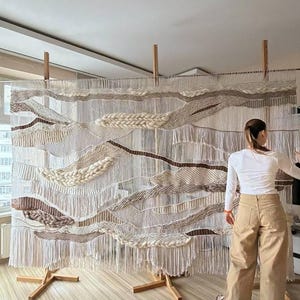 May include: A large, woven wall hanging with a textured, layered design in shades of white, beige, and brown. The artwork is mounted on a wooden frame with a fringe at the bottom. The piece is displayed in a room with a light-colored floor.