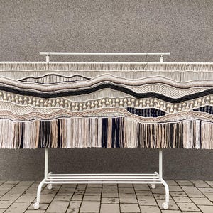 Peut inclure: Une grande tapisserie murale en macramé avec un motif ondulé en blanc, beige et noir. La tapisserie est faite de fil épais et a une frange en bas.
