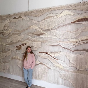 May include: A large, beige macrame wall hanging with a wavy, abstract design. The wall hanging is made with natural fibers and has a fringe border. A person is standing in front of the wall hanging.