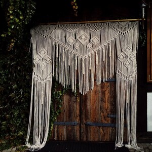 May include: A beige macrame wall hanging with geometric diamond patterns and long fringe. The handmade textile is suspended from a wooden rod, creating a bohemian decorative element. The macrame hangs in front of a wooden door.