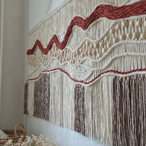 May include: A large macrame wall hanging featuring cream-colored cord, terracotta-colored wavy lines, and natural wooden beads. The lower section consists of alternating vertical panels of cream and brown fringe hanging from a horizontal wooden dowel.