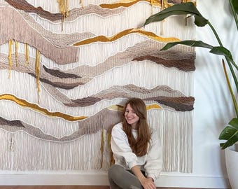 May include: A woman sits in front of a large wall hanging made of yarn. The wall hanging is a tapestry of beige, brown, and yellow yarn, creating a textured landscape design.