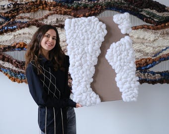 May include: A woman holding a white, textured, abstract art piece. The artwork is mounted on a brown background. A large woven wall hanging with brown, blue, and white tones is behind the woman.