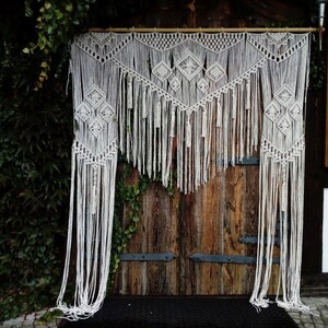 May include: A white macrame wall hanging featuring geometric diamond designs and long fringe. The handmade textile is suspended from a wooden rod, creating a bohemian decorative element. The macrame hangs in front of a rustic wooden door.
