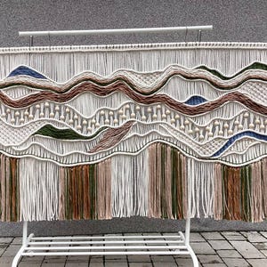 May include: A large macrame wall hanging with a landscape design. The artwork features a cream-colored background with woven patterns and accents in shades of brown, green, and blue. Wooden beads are incorporated throughout the design, and the bottom has long fringe.