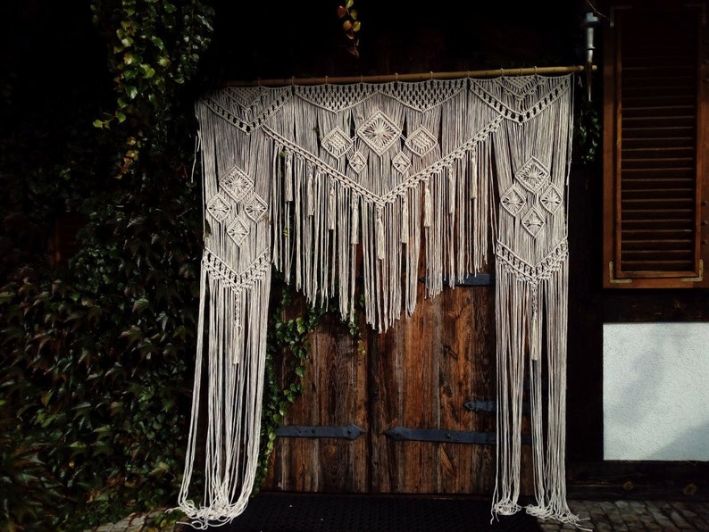 May include: A beige macrame wall hanging with geometric diamond patterns and long fringe. The handmade textile is suspended from a wooden rod, creating a bohemian decorative element. The macrame hangs in front of a wooden door.
