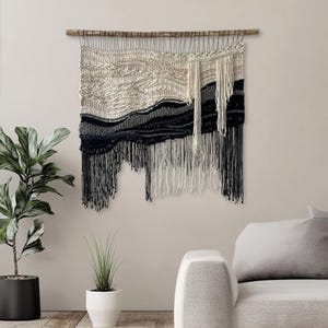 Peut inclure: Une tenture murale tissée avec une baguette en bois naturel. Le textile présente un motif texturé dans les tons crème, gris et noir, avec de longues franges en bas. Cette pièce ajoute une touche de style bohème à tout espace.
