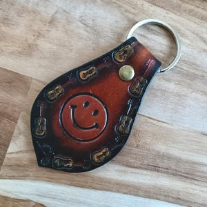May include: A teardrop-shaped brown leather keyring featuring a stamped smiley face in the centre, surrounded by a border of small guitar motifs. The leather has a dark gradient finish and is secured with a brass-coloured metal snap and ring.