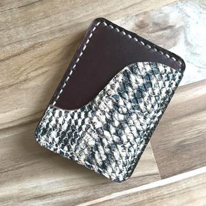 May include: A rectangular card holder featuring a combination of smooth dark brown leather and a textured black and white snakeskin pattern. The accessory is constructed with visible white stitching along the edges and a curved pocket design.