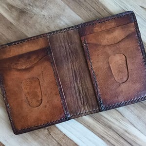 May include: Open, brown leather wallet with multiple card slots. The wallet has a worn, distressed appearance with darker edges and visible stitching. The interior features a lighter brown colour, and the wallet is open, displaying its compartments.