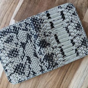May include: A rectangular wallet featuring a textured, scale-like pattern in shades of black, white, and light grey. The item has rounded corners and visible stitching along the edges, displaying a consistent, repeating organic design throughout.