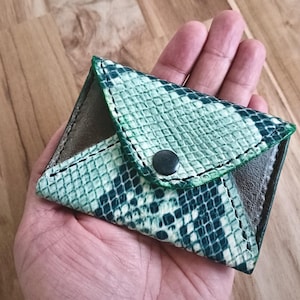 May include: A small envelope-style wallet featuring a textured green and white snakeskin pattern on the exterior flap. The wallet is constructed with dark green stitching along the edges and includes a single black circular snap button closure.