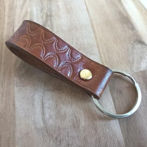May include: A brown leather keychain with a decorative embossed pattern. The keychain features a silver metal ring and a gold-colored rivet. The leather strap is approximately 4 inches long.
