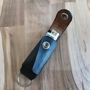 May include: A black leather lighter case with a silver metal snap button closure and a silver metal keyring attached at the bottom. The case is partially open, revealing a white and silver lighter inside, with the interior of the leather flap visible.