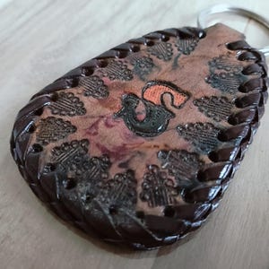 Op de afbeelding: Een druppelvormige leren sleutelhanger met een donkerbruine gevlochten rand en een lichtbruin middenstuk met een gestempelde zwarte en oranje letter S. Het ontwerp bevat een herhalend patroon van kleine, gestempelde boommotieven rondom.