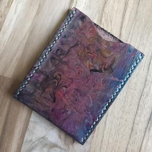 May include: A rectangular leather card holder featuring a marbled pattern of deep purple, red, and blue tones. The edges are secured with thick, light blue stitching, and the top edge has a curved cutout for easy access to cards.