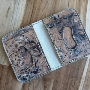 May include: A bi-fold card holder featuring a marbled pattern in shades of tan, black, and white. The exterior panels have oval-shaped cutouts for easy card access, and the interior spine is constructed from a solid white material with visible stitching.