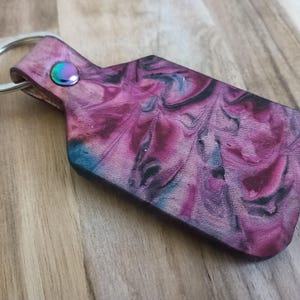 May include: A rectangular keyring with rounded corners featuring a marbled pattern of deep magenta, black, and teal hues. The keyring is attached to a silver-toned metal split ring by a small loop secured with a single iridescent metal rivet.