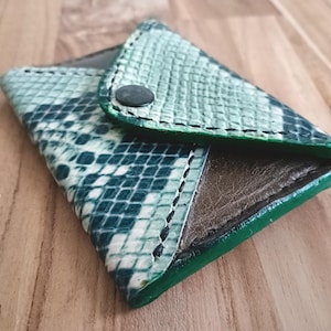 May include: A small envelope-style wallet featuring a textured teal and white snakeskin pattern on the exterior flap. The wallet is constructed with dark brown leather panels, visible black stitching along the edges, and a single black snap button.