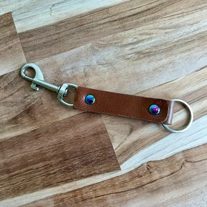 May include: A brown leather keychain strap with a silver-colored clasp and key ring. The strap is secured with two iridescent rivets. The keychain is designed for attaching keys or other small items.