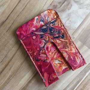 May include: A rectangular leather card holder featuring a vibrant marbled pattern in shades of magenta, deep blue, and cream. The accessory has visible stitching along the edges and a central cutout slot on the front for easy access to cards.